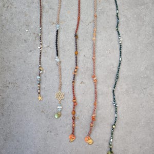 May include: Five handmade hair wraps with various bead and charm designs. The wraps feature a range of colors, including brown, black, red, and green, with gold-colored charms and star and flower pendants. Each wrap is unique, with different bead sizes and shapes.