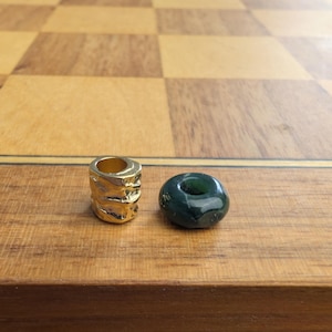 May include: A cylindrical gold-toned metal bead with a textured, irregular surface alongside a smooth, dark green circular stone bead with a central hole. Both items are small, decorative components intended for jewelry making or crafting projects.