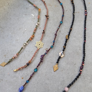 May include: Five handmade hair wraps with various colors and designs. Each wrap features a braided cord adorned with beads, crystals, and charms. The wraps include gold-colored charms, star and flower pendants, and a feather charm. The color palette includes green, brown, black, and purple.