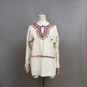 May include: A white cotton tunic with colorful embroidery around the neckline and cuffs. The embroidery features a geometric pattern with tassels.