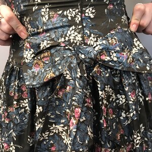 Puede incluir: Un vestido verde oscuro con estampado floral en tonos azules, blancos y rosas. El vestido tiene un lazo en la cintura y una falda de capas.