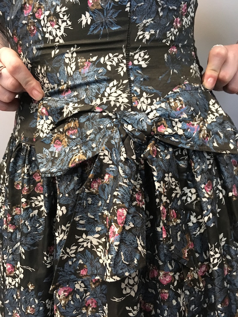 Puede incluir: Un vestido verde oscuro con estampado floral en tonos azules, blancos y rosas. El vestido tiene un lazo en la cintura y una falda de capas.