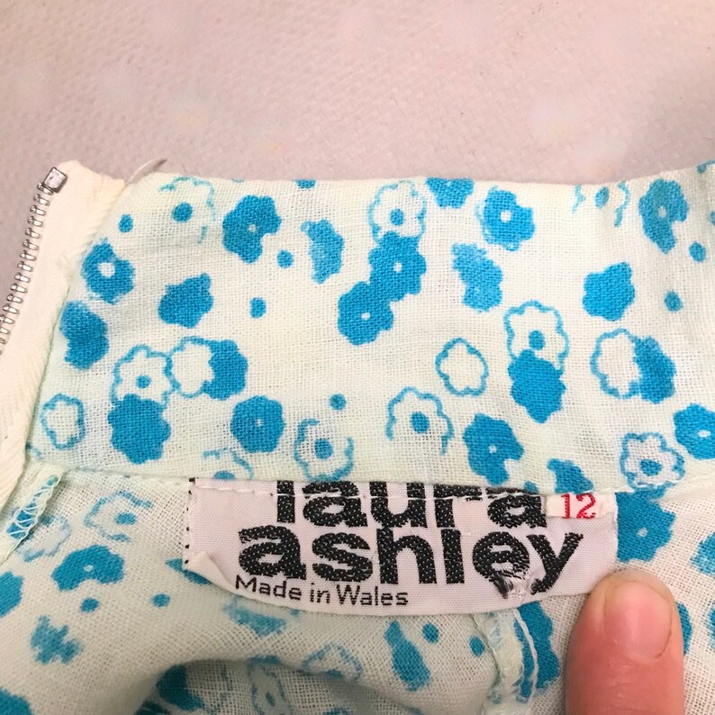 May include: A white dress with a blue floral pattern and a black label that reads "laura ashley" and "Made in Wales". The label also has a size tag that says "12".