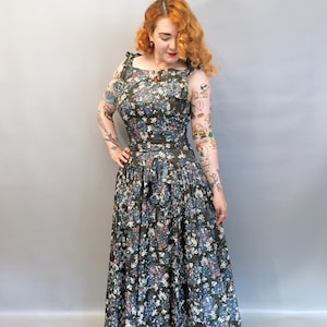 Puede incluir: Un vestido largo y fluido de estampado floral con un corpi&ntilde;o ajustado y una falda completa. El vestido tiene un fondo verde oscuro con flores rosas, azules y blancas. El vestido tiene un escote cuadrado y tirantes finos.