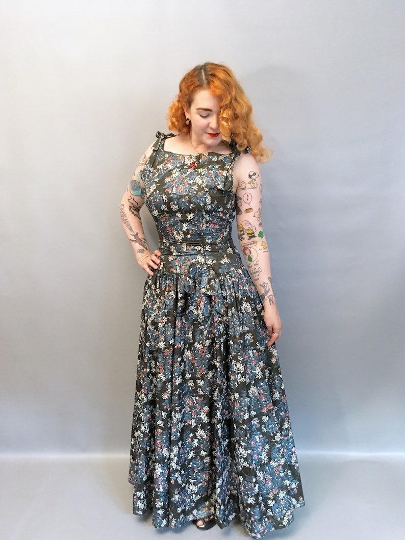 Puede incluir: Un vestido largo y fluido de estampado floral con un corpi&ntilde;o ajustado y una falda completa. El vestido tiene un fondo verde oscuro con flores rosas, azules y blancas. El vestido tiene un escote cuadrado y tirantes finos.