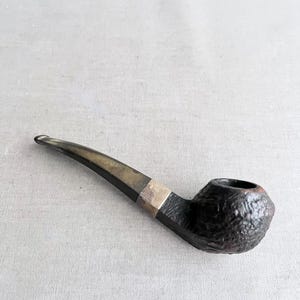 Pode incluir: Um cachimbo de tabaco vintage com uma tigela escura e texturizada e um bocal curvo. A haste é de cor escura com uma faixa prateada perto da tigela. O cachimbo repousa sobre uma superfície cinza claro.