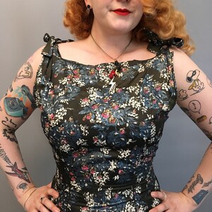 Puede incluir: Una mujer lleva un vestido de estampado floral verde con escote cuadrado y tirantes de lazo. El vestido tiene un corpi&ntilde;o ajustado y una falda completa. La mujer tiene el pelo rojo y lleva l&aacute;piz labial rojo.