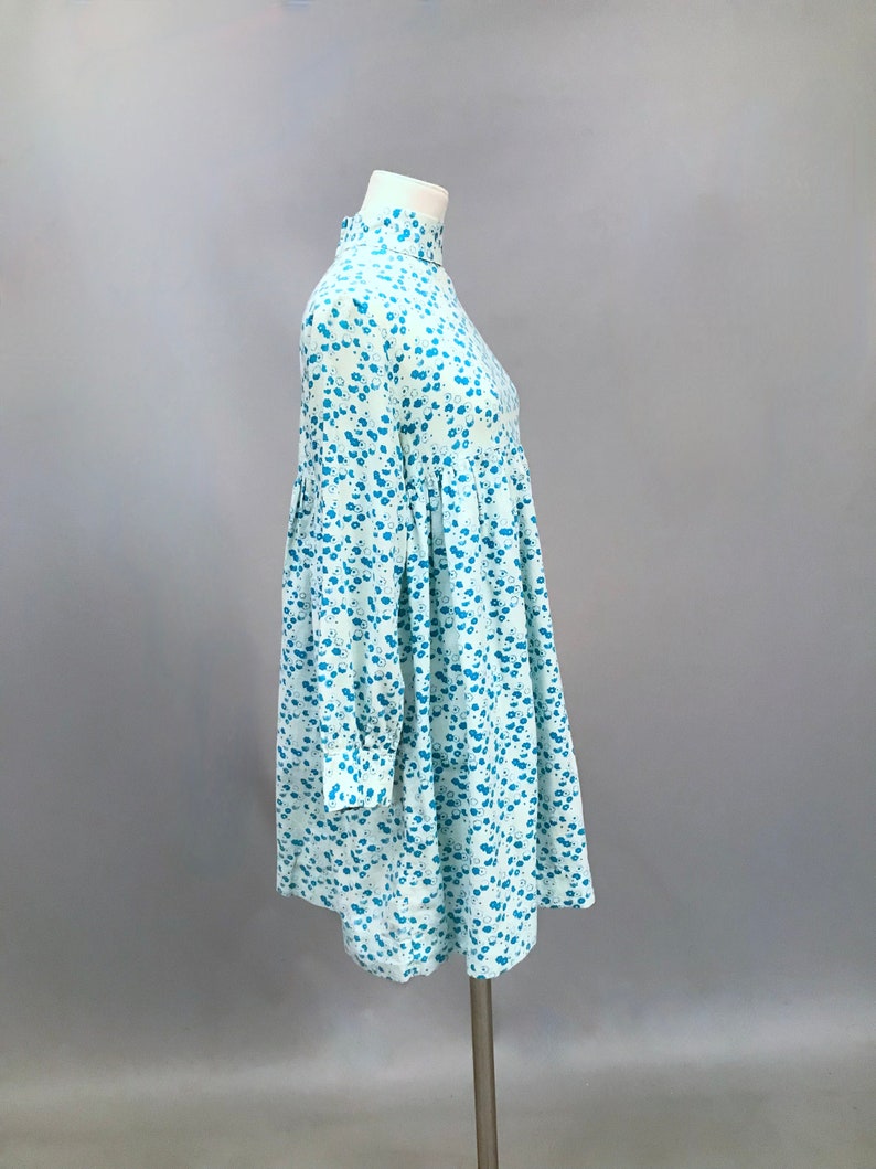 May include: A light blue dress with a white and blue floral pattern. The dress has a high neck and long sleeves. It is fitted at the waist and flows out to a slightly flared skirt.