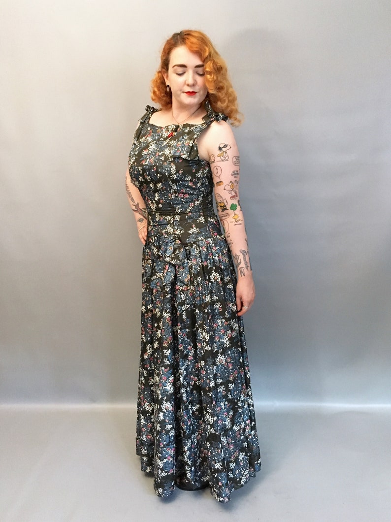 Puede incluir: Un vestido largo y fluido con estampado floral, con un corpi&ntilde;o ajustado y una falda completa. El vestido tiene un fondo azul oscuro con flores blancas y rosas. El vestido tiene un lazo en la cintura y un escote con volantes.