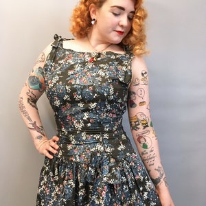 Puede incluir: Una mujer con un vestido de estampado floral verde y azul con cuello redondo y tirantes. El vestido tiene una falda con volantes y la mujer tiene el pelo rojo y tatuajes en los brazos.