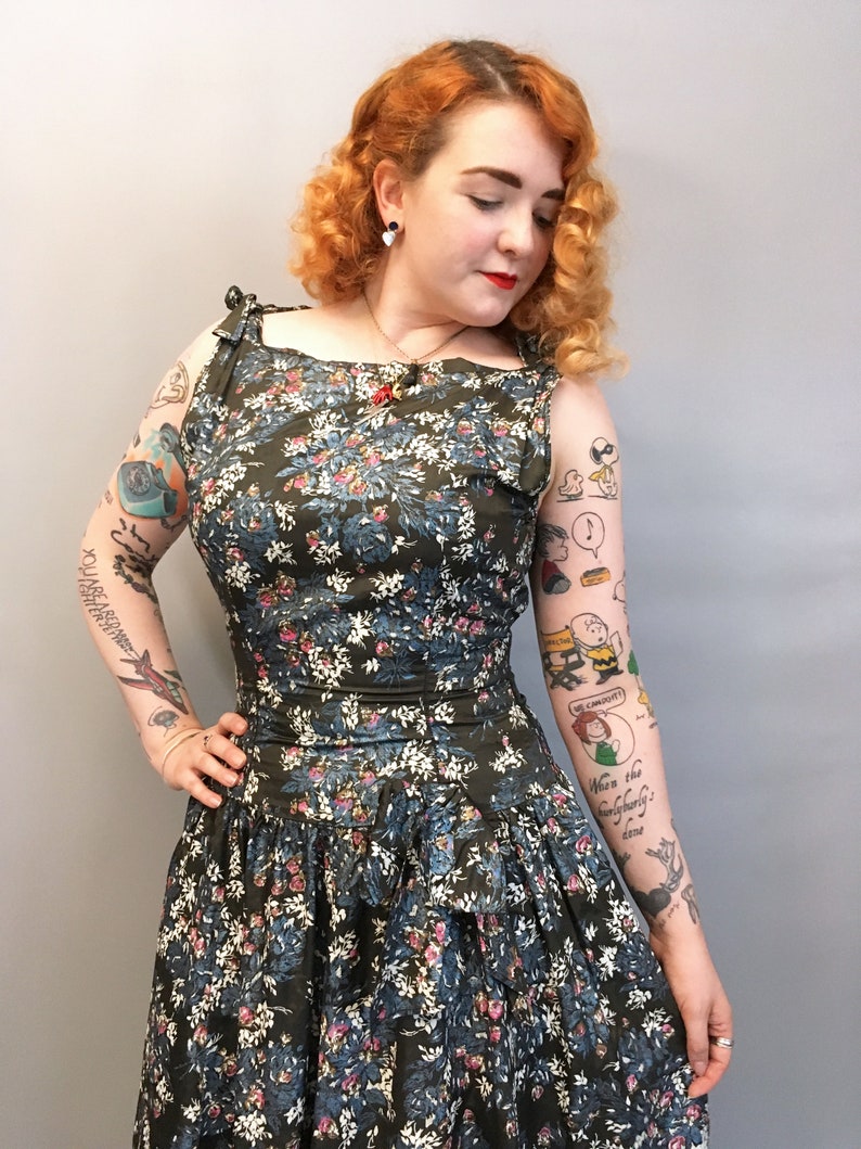 Puede incluir: Una mujer con un vestido de estampado floral verde y azul con cuello redondo y tirantes. El vestido tiene una falda con volantes y la mujer tiene el pelo rojo y tatuajes en los brazos.