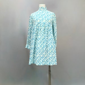 May include: A white dress with a blue floral pattern. The dress has a high neck and long sleeves. It is a simple, elegant style.