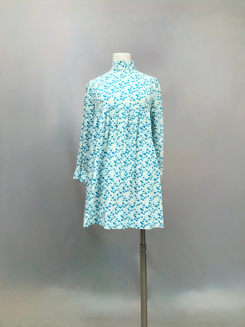 May include: A white dress with a blue floral pattern. The dress has a high neck and long sleeves. It is a simple, elegant style.