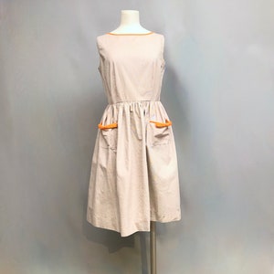 以下が含まれることがあります： 薄いグレーと白のストライプ柄のノースリーブドレス。ドレスはフィットしたボディとフルスカート。ネックラインとポケットは明るいオレンジの生地でトリミングされています。