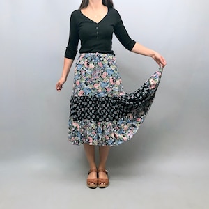 Op de afbeelding: Een zwart shirt met lange mouwen met een rok met bloemenprint. De rok heeft drie lagen en is een mix van zwart, blauw, roze en groen.