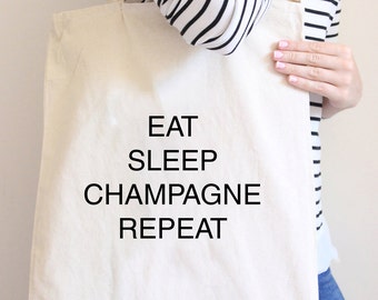 May include: A white canvas tote bag with black text that reads "Eat Sleep Champagne Repeat".