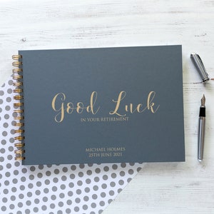 May include: A gray hardcover guest book with a gold spiral binding. The cover has the text "Good Luck in Your Retirement" and "Michael Holmes 25th June 2021" in gold lettering.
