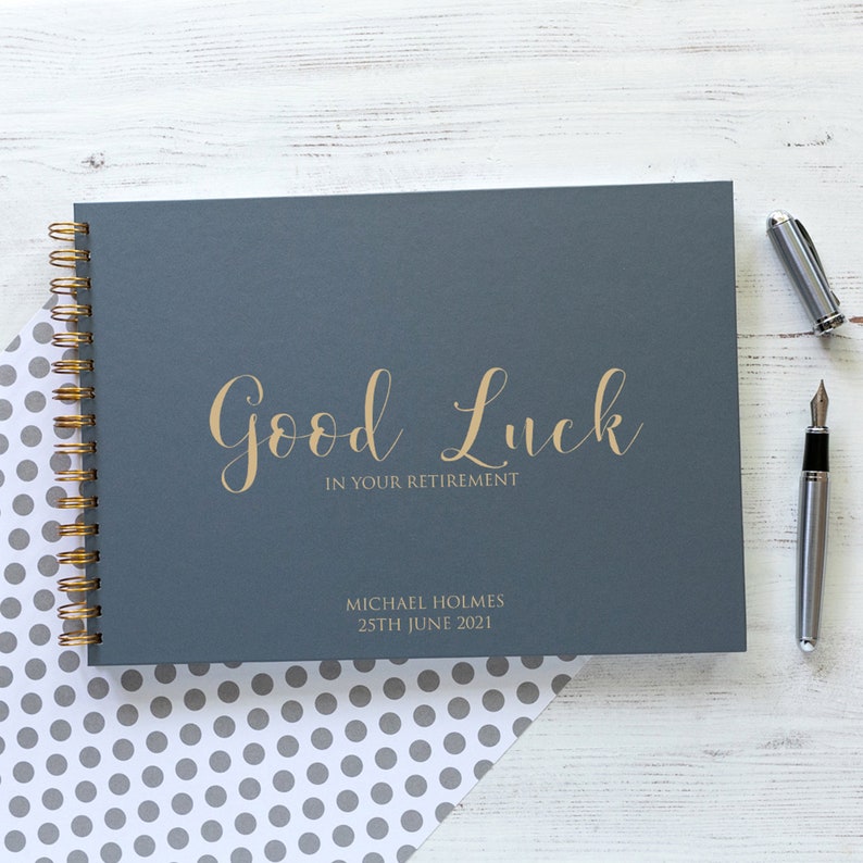 May include: A gray hardcover guest book with a gold spiral binding. The cover has the text "Good Luck in Your Retirement" and "Michael Holmes 25th June 2021" in gold lettering.