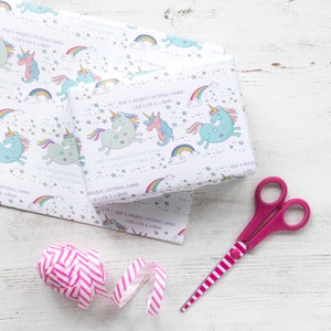 May include: White wrapping paper with a repeating pattern of rainbows, stars, and unicorns. The paper has the text "Have a magical Christmas Emma Love Kate & William" printed on it. A pink and white striped ribbon and pink scissors are also in the image.