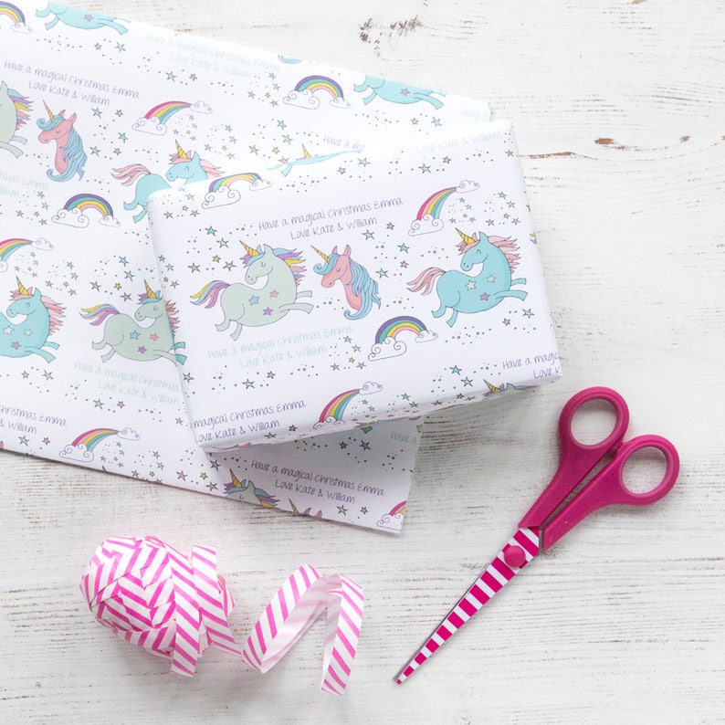 May include: White wrapping paper with a repeating pattern of rainbows, stars, and unicorns. The paper has the text "Have a magical Christmas Emma Love Kate & William" printed on it. A pink and white striped ribbon and pink scissors are also in the image.