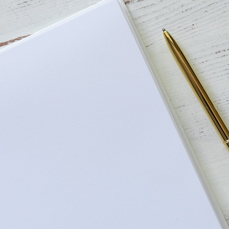 May include: A blank white notebook with a gold pen lying on a white wooden surface.