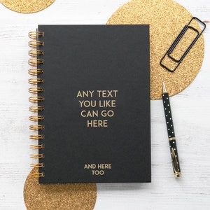 May include: A black spiral bound notebook with gold accents. The notebook has the text "ANY TEXT YOU LIKE CAN GO HERE AND HERE TOO" printed on the front cover.