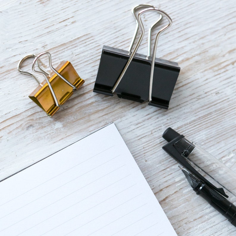 May include: A white lined notebook with a black pen and two binder clips, one gold and one black, on a white wooden surface.