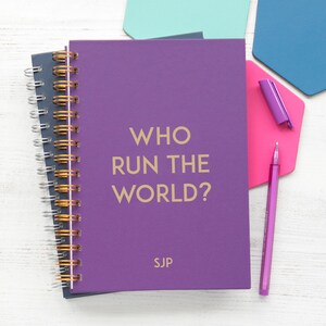 May include: Purple spiral bound notebook with the text "WHO RUN THE WORLD?" in gold lettering and the initials "SJP" in gold lettering at the bottom. The notebook is on a white wooden surface with a pink and blue hexagon shaped paper.