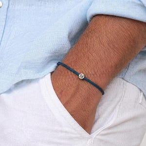 May include: A dark blue bracelet with a small, round silver charm featuring the letter "M". The bracelet is made of a woven cord and is worn on the wrist. The person is wearing a light blue shirt and white pants.