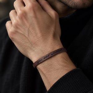 Puede incluir: Una pulsera fina de cuero marrón oscuro con el texto grabado 'you ever wonder' en una fuente cursiva. La pulsera se lleva alrededor de la muñeca y presenta un acabado liso y mate con un diseño de banda estrecha y sencilla.
