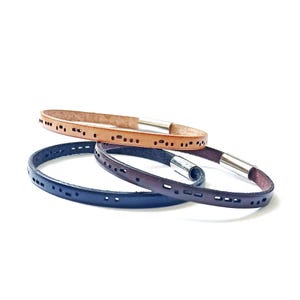 Puede incluir: Tres pulseras de cuero en tonos beige, azul marino y marrón, cada una con una serie de pequeños agujeros espaciados uniformemente. Las pulseras tienen un diseño minimalista con cierres de metal plateado. Un accesorio elegante.
