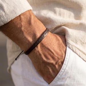 Può includere: Braccialetto in pelle marrone scuro con la scritta incisa "end of You always". Il braccialetto è indossato al polso, con chiusura a coulisse. La persona indossa una camicia di lino colorata e pantaloni bianchi.