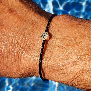 May include: A black cord bracelet with a small, round, silver-coloured charm. The charm has a gold-coloured band and a detailed design. The bracelet is worn on a person's wrist, with a blue water background.
