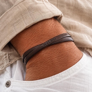 May include: A dark brown leather bracelet with the engraved date "26.03.2004". The bracelet wraps around the wrist. The person is wearing a light beige button-down shirt and a white shirt.