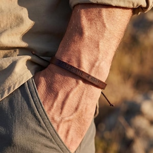 Puede incluir: Una pulsera de cuero marrón con las coordenadas "37.12306°N, -4.65611°W" grabadas. La pulsera se cierra con un cordón. La persona lleva una camisa color beige y pantalones grises.