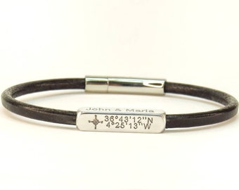 Puede incluir: Una pulsera de cuero marrón oscuro con cierre plateado y placa rectangular plateada. La placa está grabada con los nombres "John & Maria", una rosa de los vientos y coordenadas: 36°43'12"N 4°25'13"W.