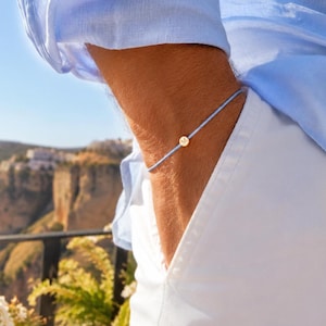 Puede incluir: Una pulsera de cordón azul claro con un pequeño dije circular de color dorado. La pulsera se lleva en la muñeca. La persona lleva una camisa azul claro y pantalones blancos.