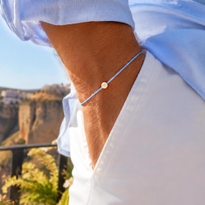 Peut inclure: Un bracelet bleu clair avec un petit charme circulaire de couleur or. Le charme porte la lettre "M" gravée. Le bracelet est porté au poignet, avec une chemise bleu clair et un pantalon blanc en arrière-plan.