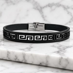 Peut inclure: Bracelet en cuir noir avec un motif géométrique de clé grecque. Le bracelet est doté d'un fermoir en acier inoxydable et est présenté sur une surface en marbre. Le design est découpé, créant un accessoire élégant. Circonférence d'environ 20 cm.
