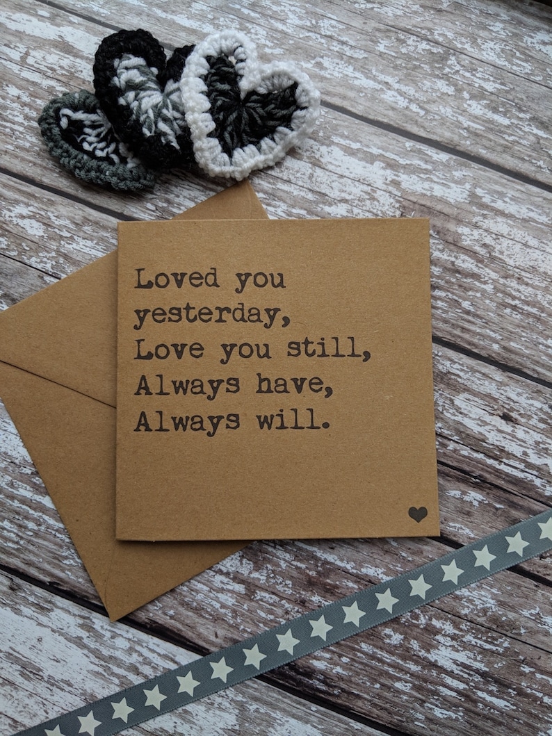 Puede incluir: Una tarjeta marr&oacute;n con el texto "Loved you yesterday, Love you still, Always have, Always will." La tarjeta est&aacute; sobre una superficie de madera con una cinta gris con estrellas blancas y tres corazones de ganchillo en blanco y negro.