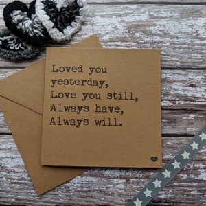 Puede incluir: Una tarjeta marr&oacute;n con el texto "Loved you yesterday, Love you still, Always have, Always will." La tarjeta est&aacute; sobre una superficie de madera con una cinta gris con estrellas blancas y dos corazones de ganchillo.