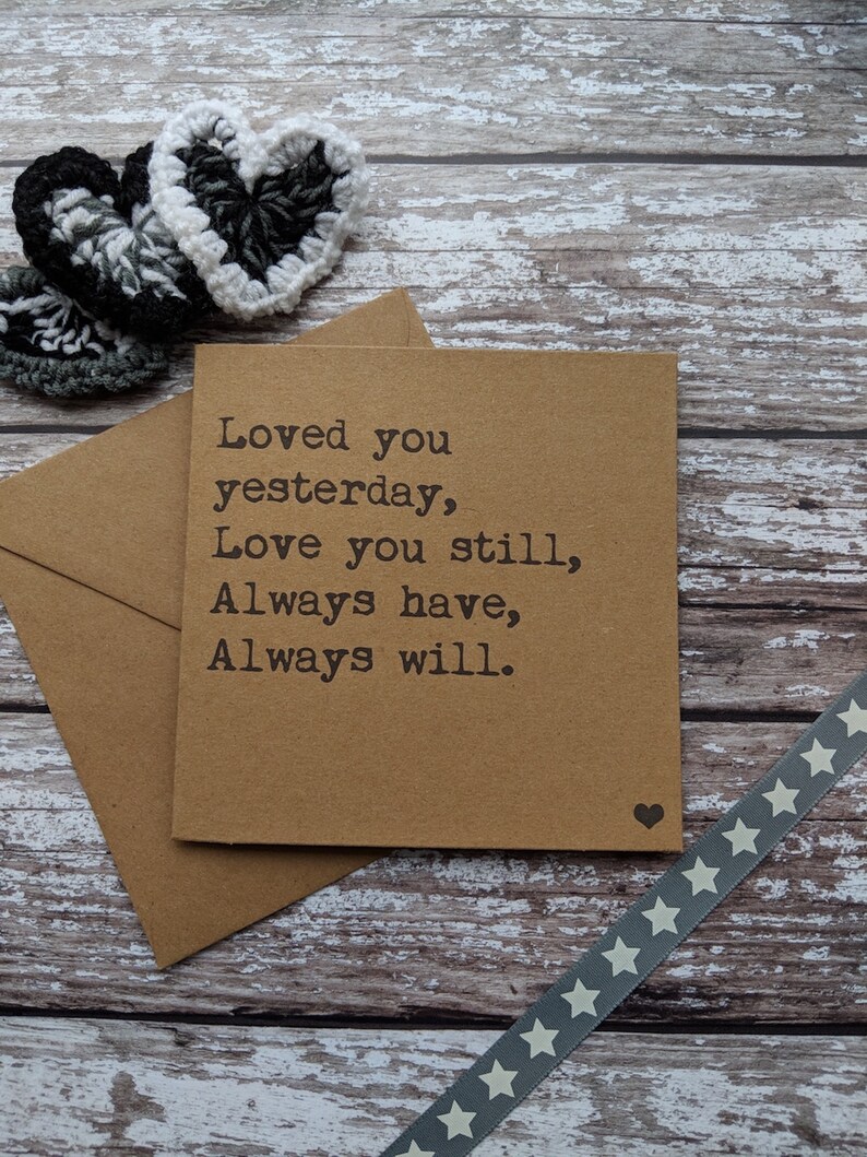 Puede incluir: Una tarjeta marr&oacute;n con el texto "Loved you yesterday, Love you still, Always have, Always will." La tarjeta est&aacute; sobre una superficie de madera con una cinta gris con estrellas blancas y dos corazones de ganchillo.