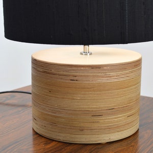 Puede incluir: Una pantalla de l&aacute;mpara negra se asienta sobre una base de l&aacute;mpara de madera cil&iacute;ndrica hecha de madera contrachapada apilada.