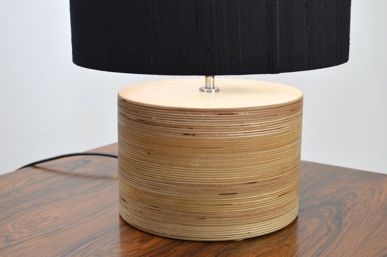 Puede incluir: Una pantalla de l&aacute;mpara negra se asienta sobre una base de l&aacute;mpara de madera cil&iacute;ndrica hecha de madera contrachapada apilada.