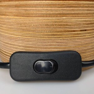Puede incluir: Primer plano de un interruptor de encendido/apagado negro conectado a un cable negro. El cable est&aacute; envuelto alrededor de una base de l&aacute;mpara de madera hecha de finas capas de madera.