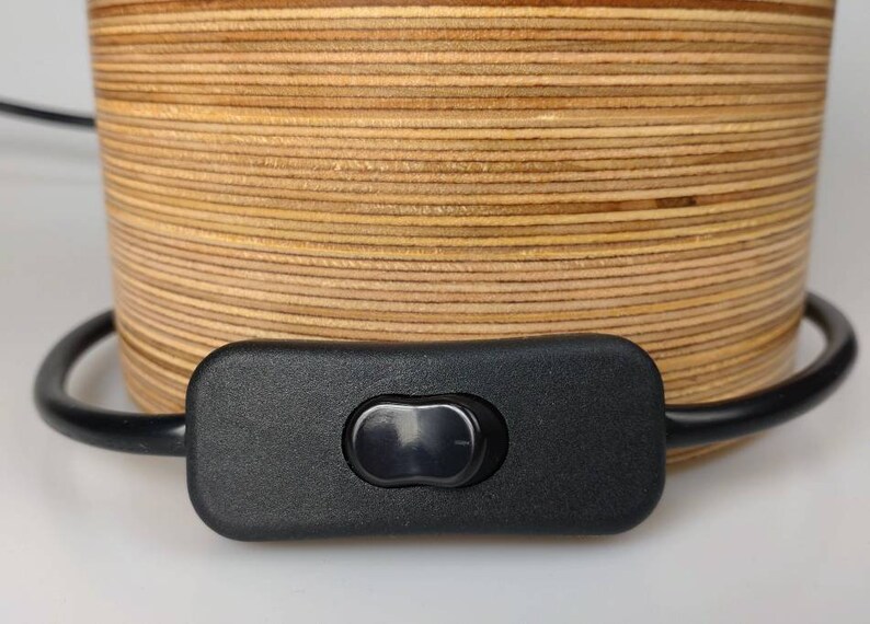 Puede incluir: Primer plano de un interruptor de encendido/apagado negro conectado a un cable negro. El cable est&aacute; envuelto alrededor de una base de l&aacute;mpara de madera hecha de finas capas de madera.