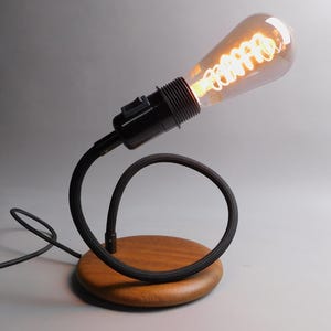 Könnte beinhalten: Eine schwarze Schreibtischlampe mit einem flexiblen Kabel und einem Holzsockel. Die Lampe hat eine Glühbirne im Vintage-Stil mit einem spiralförmigen Filament.