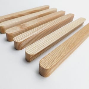 Könnte beinhalten: Sechs hellfarbige Holzstäbe mit abgerundeten Enden sind diagonal auf einer weißen Oberfläche angeordnet. Die Holzmaserung ist sichtbar, und die Stäbe haben eine glatte, natürliche Oberfläche. Die Stäbe sind wahrscheinlich für ein Dekorations- oder Bastelprojekt.