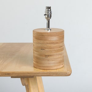 Puede incluir: Una l&aacute;mpara de mesa de madera con una base cil&iacute;ndrica hecha de madera contrachapada apilada. La l&aacute;mpara tiene un casquillo de metal plateado y un cable blanco.