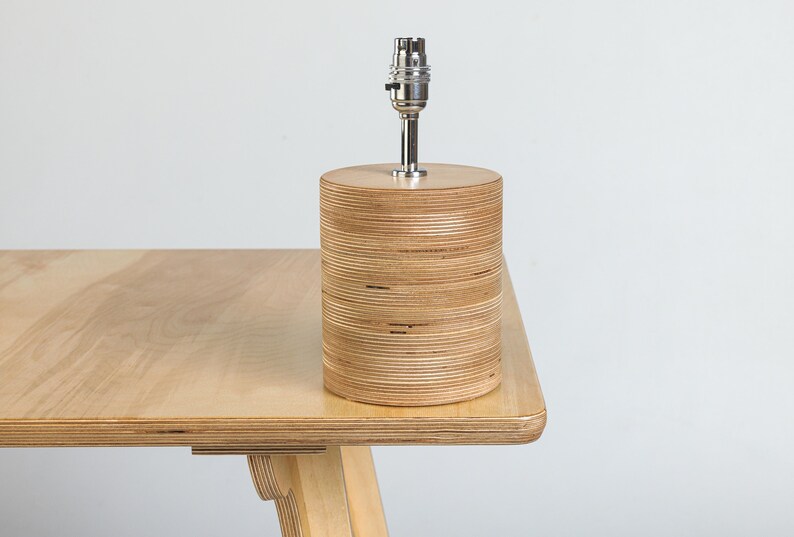 Puede incluir: Una l&aacute;mpara de mesa de madera con una base cil&iacute;ndrica hecha de madera contrachapada apilada. La l&aacute;mpara tiene un casquillo de metal plateado y un cable blanco.